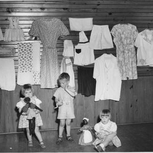 Pleasant Hills Home Demonstration Club project, Edgecombe County, North Carolina. Children wearing clothing and holding toys, and a display of clothing hanging on the wall behind them, all made from feed sacks.