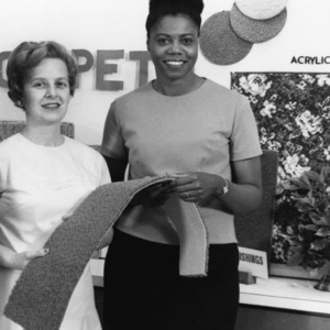 Two women holding carpet sample, carpet display in background, May 24, 1967