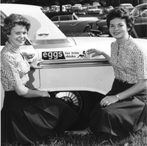 Two young women, 4-H team demonstration members from Chatham County, North Carolina, posing by a bumper sticker on a car that reads, "Eggs for trim chicks."