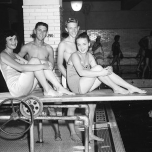 Two girls in swimsuits sitting on diving board, with two boys behind them, during North Carolina State 4-H Club Week