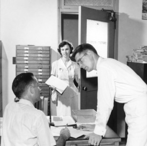 Two men talking in the Department of Information office as woman enters, during North Carolina State 4-H Club Week