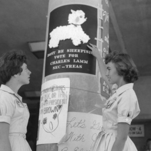 Two 4-H club girls considering an election poster reading "Don't be sheepish, vote for Charles Lamm, sec-treas," during North Carolina State 4-H Club Week