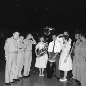 4-H club girl playing tuba while other 4-H members and men in army uniforms plug their ears, during North Carolina State 4-H Club Week
