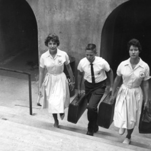 Two 4-H club girls and one 4-H club boy, with luggage in hand, walking up the stairs from the pedestrian tunnel underneath the railroad tracks at North Carolina State College, during North Carolina State 4-H Club Week