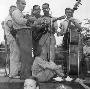 Five unidentified men playing string instruments, including mandolin, guitar, and bass for a group of 4-H club members