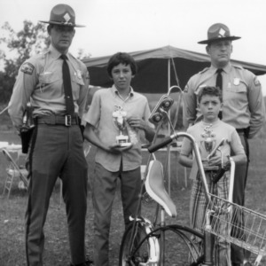 Two 4-H club boys holding trophies and standing between North Carolina highway patrol officers at the Asheville bicycle rodeo