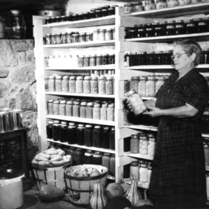 Unidentified woman standing in front of shelves of jars, holding jar of preserved fruit