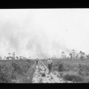 Fire in the bay: Smoke rises in background as two men walk towards fire on sandy track in foreground