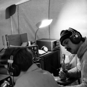 Two men sitting in front of electronic equipment and talking into microphones