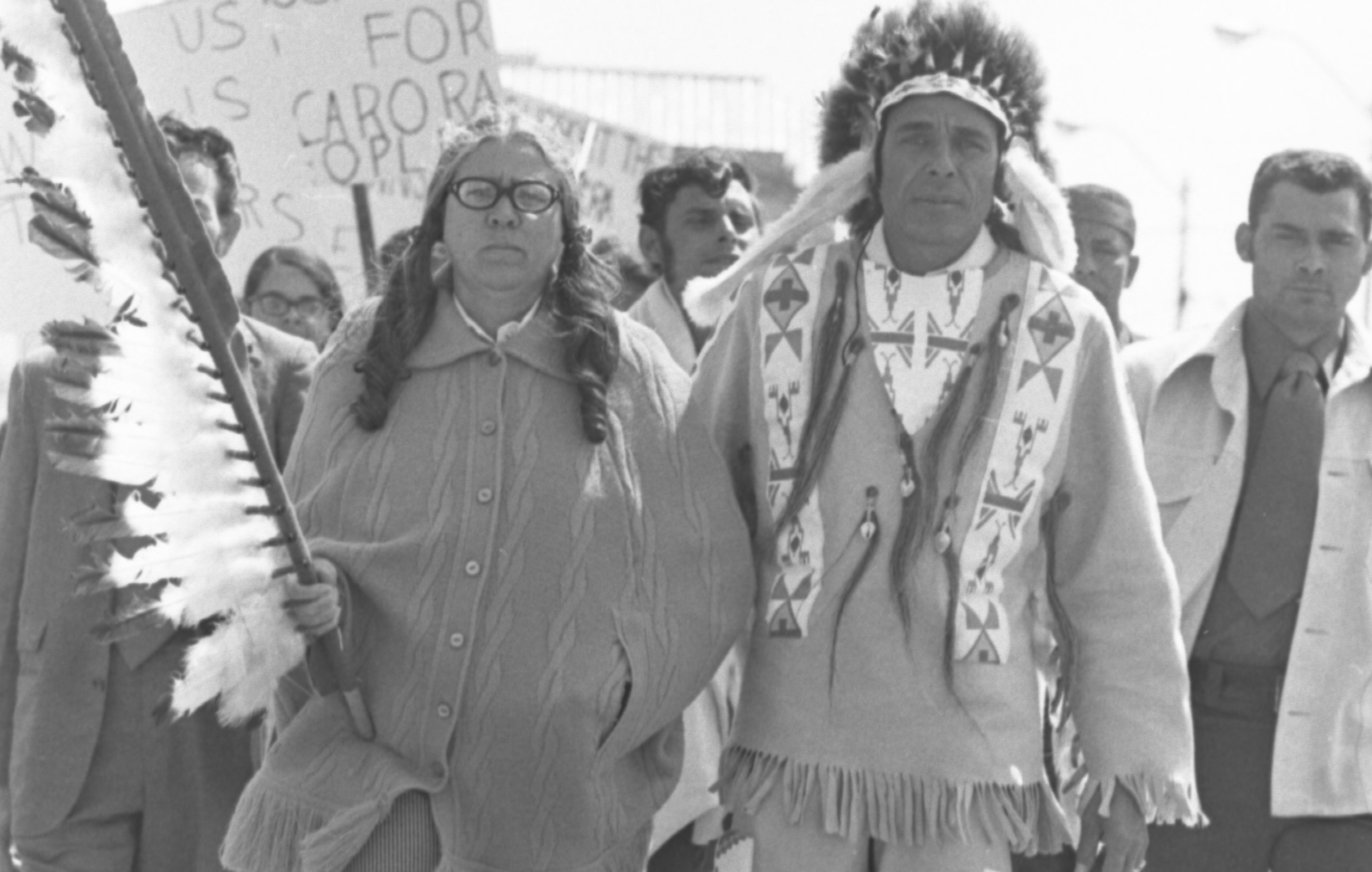 Two people in traditional dress march in front of people with protest signs