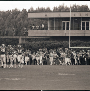 Football players, cheerleaders, and marching band on field, circa 1969-1975
