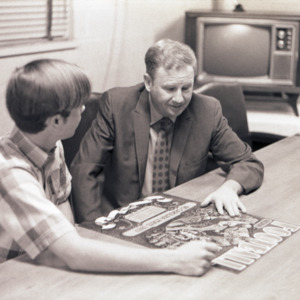 Swimming coach Willis R. Casey and other at table with poster, circa 1969 - 1975