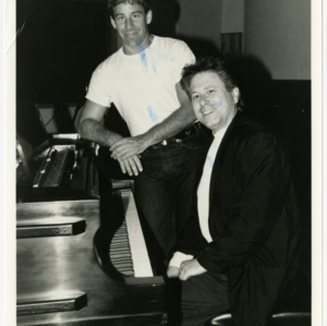 Two men smile by a piano
