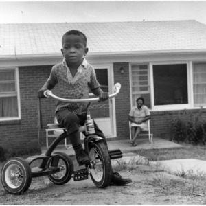 Little boy riding a tricycle while woman watches from house