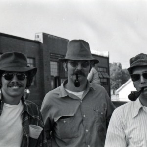 Three men in hats and sunglasses, two of them with pipes