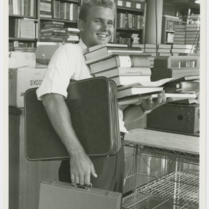 Student inside bookstore, carrying a large stack of books and suitcases