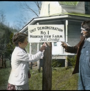 Two people in front of a house pointing to  a sign that reads, "Unit Demonstration No. 1 - Mountain View Farm - Bill Conley "