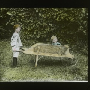Young boy wheeling younger boy in wheelbarrow, colorized, circa 1910